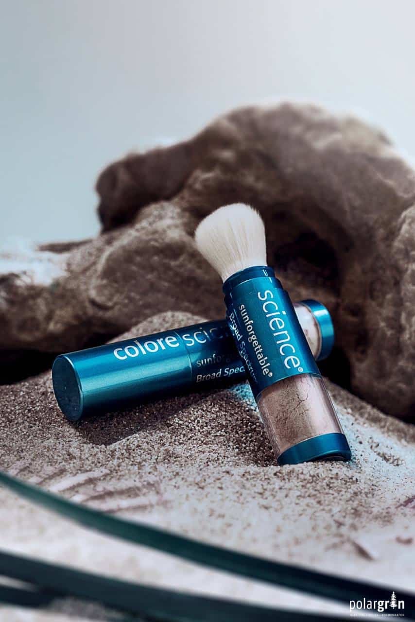 Pinsel und Puderdose von Colorescience auf Sand vor einem Stein
