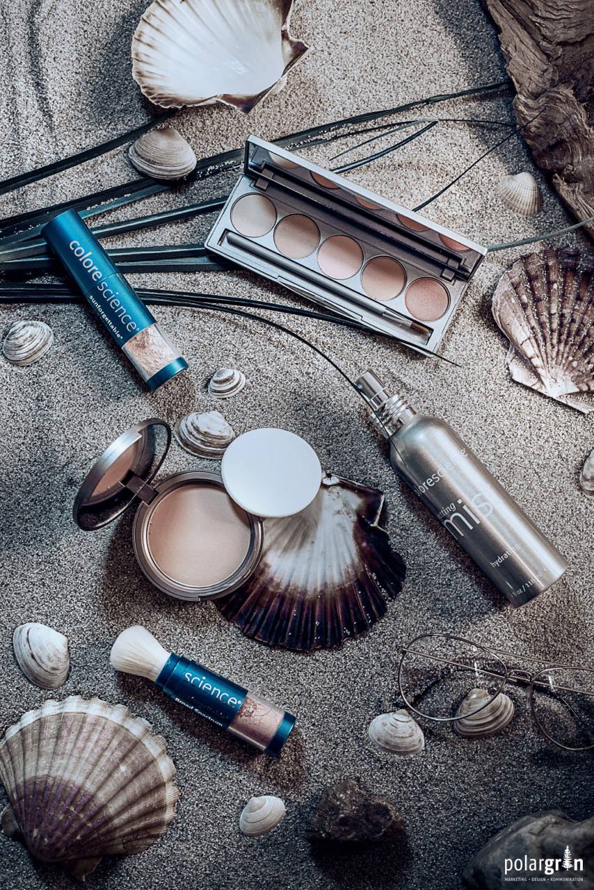 Kosmetikprodukte mit Pinsel und Lidschattenpalette auf Sand, dekoriert mit Muscheln