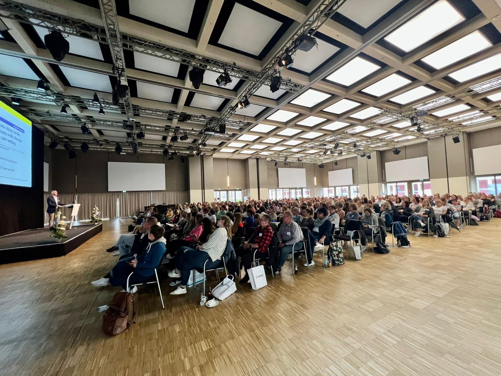 Voll besetzter Kongresssaal mit Bühne und Leinwand